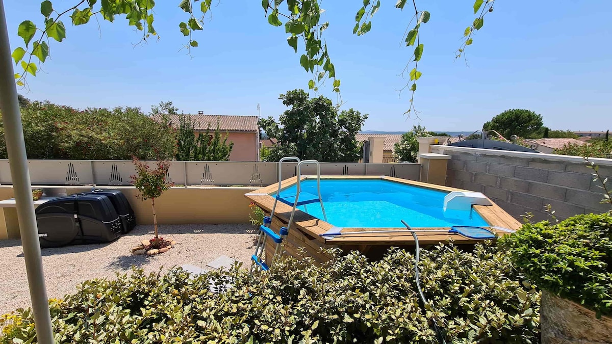 An outdoor space features a wooden above-ground pool surrounded by greenery. A ladder leads into the water, and a gravel area is visible with a few lounge chairs in the background. The scene is framed by trees and a clear blue sky.