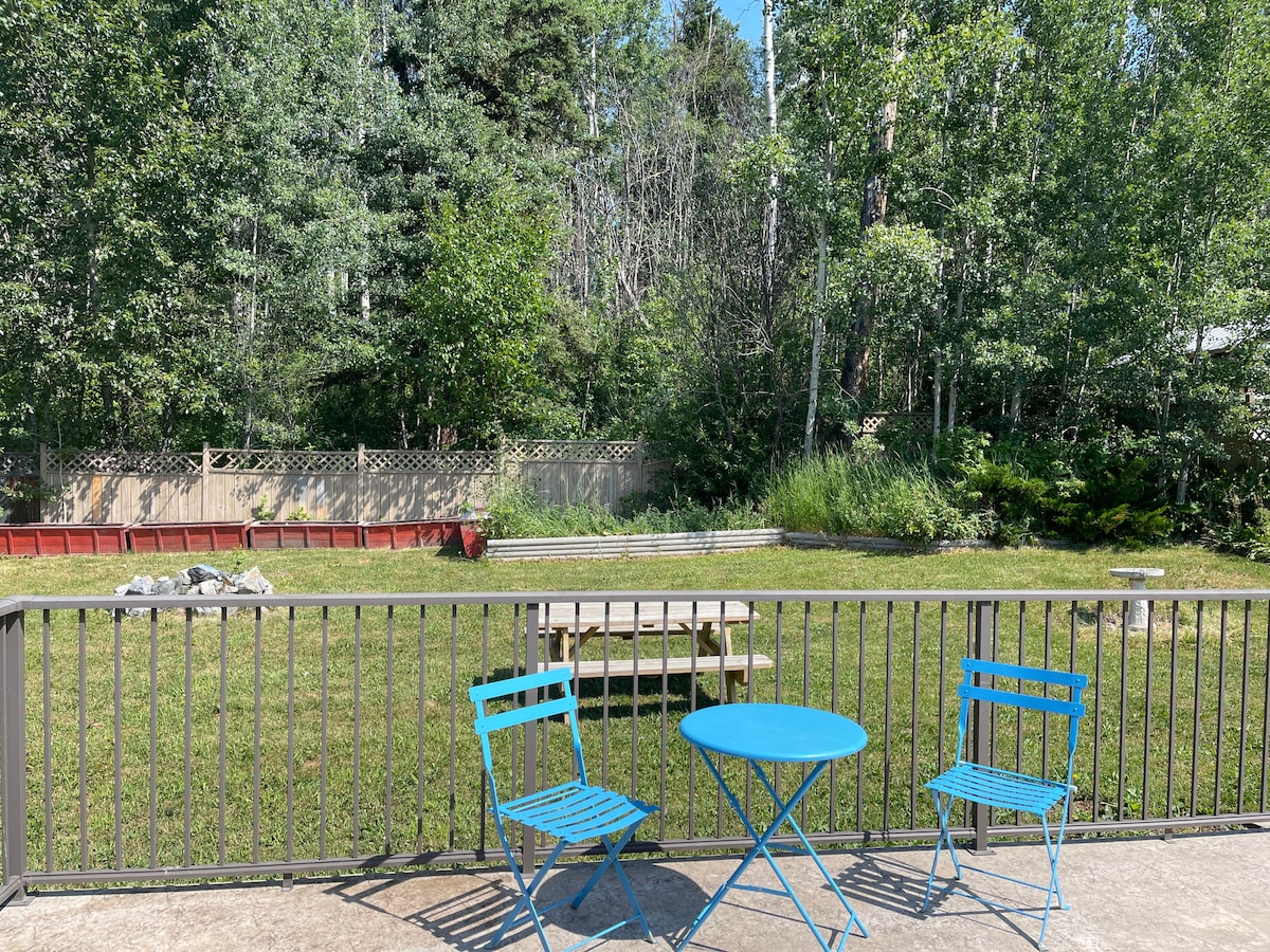 A spacious outdoor area is visible, featuring a green grassy lawn surrounded by trees. Two blue metal chairs and a small round table are positioned on a concrete patio, inviting relaxation while enjoying the natural scenery. A wooden picnic table can be seen in the distance.