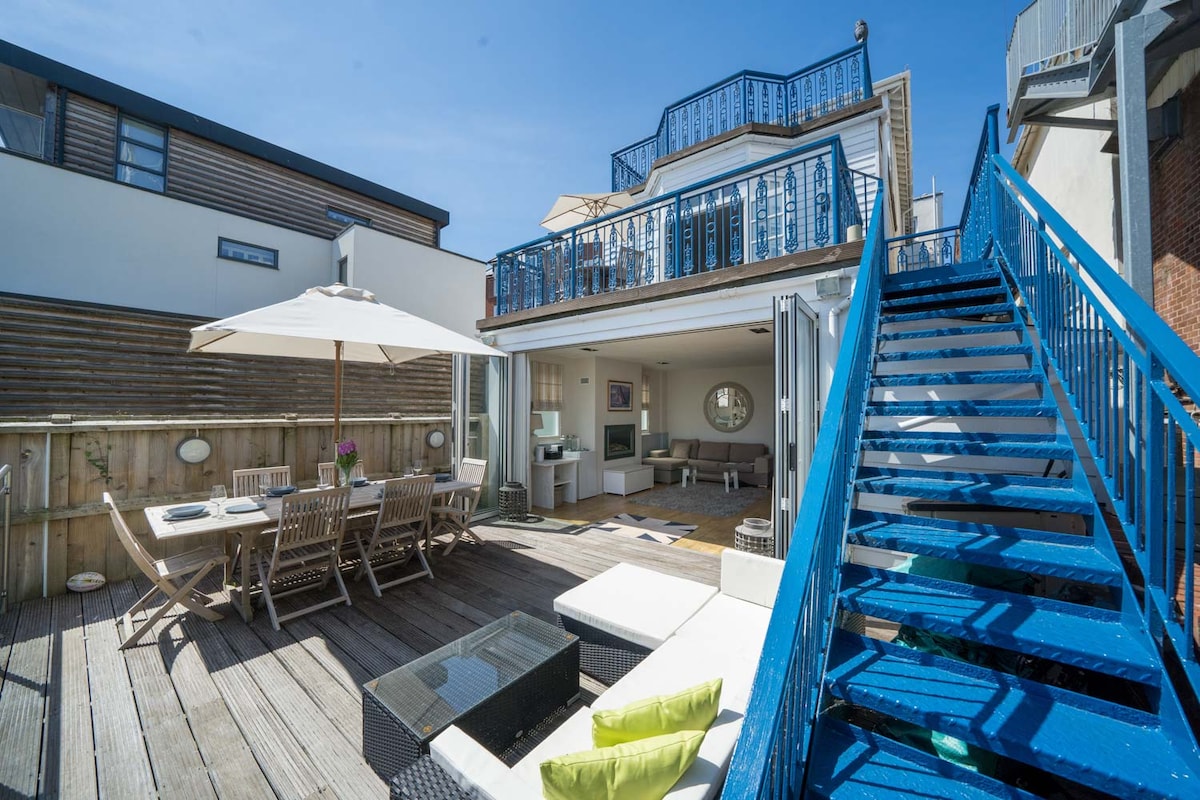 An outdoor terrace showcases a large dining table surrounded by several wooden chairs and an umbrella for shade. Bright blue stairs lead to the upper level, while a cozy seating area with cushions is visible, inviting relaxation in the sun.