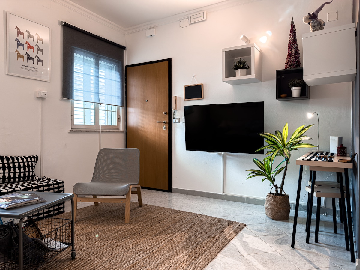 A modern living area is highlighted by a comfortable lounge chair and a stylish coffee table. A flat-screen TV is mounted on the wall, while a vibrant plant adds a touch of greenery. Natural light filters through the window, enhancing the bright ambiance.