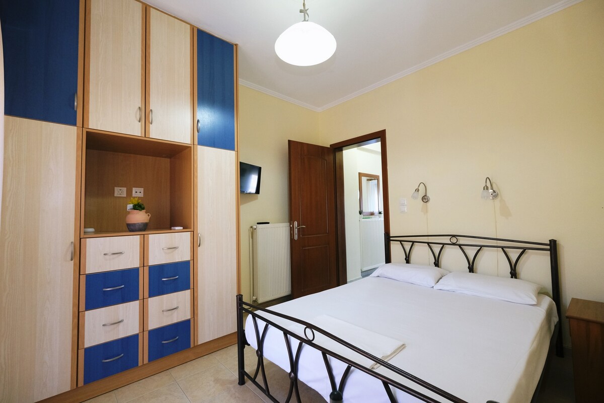 A well-organized sleeping area features a spacious bed with light bedding. A combination wardrobe with blue and beige finishes stands against one wall, while a small wooden nightstand is positioned beside the bed. A door leads to a bathroom, and wall-mounted lights offer additional illumination.
