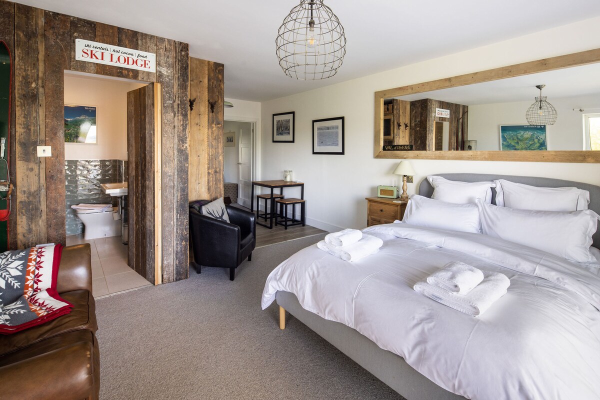 A spacious studio showcases a Kingsize bed dressed in crisp white linens, accompanied by plush towels. The room features rustic wooden accents and a large mirror, with a functional seating area equipped with a small table. An ensuite bathroom is visible, providing convenience.