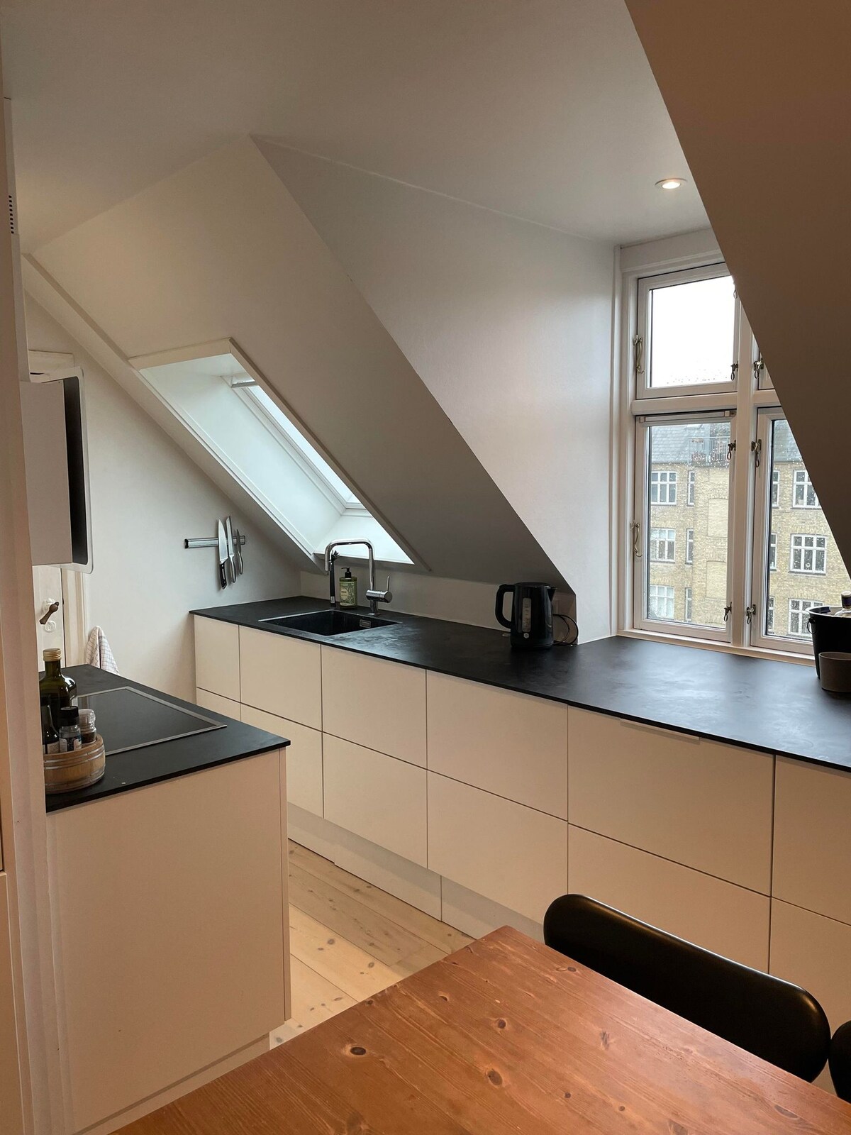 An airy kitchen features sloped ceilings and large windows allowing natural light to fill the space. The sleek countertop provides room for appliances and cooking essentials. A wooden dining table is positioned nearby, enhancing the functionality of the area.