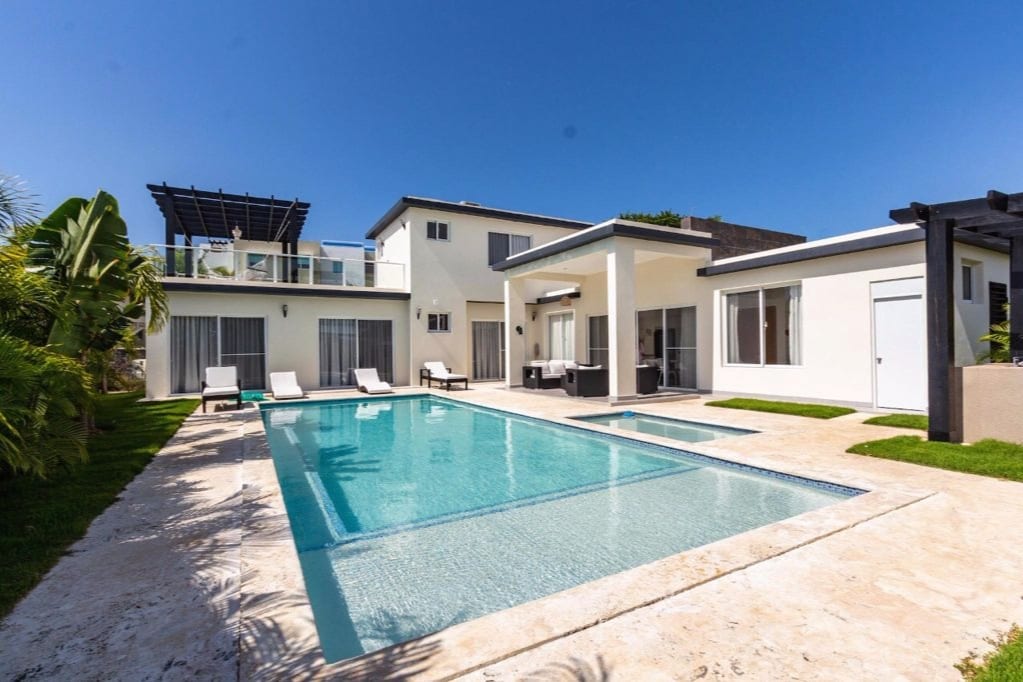 A spacious villa is set within a well-kept exterior, featuring a large private pool surrounded by lounge chairs. The building showcases modern architecture, with expansive glass doors leading to living areas, and a second-floor balcony offering views of the pool.