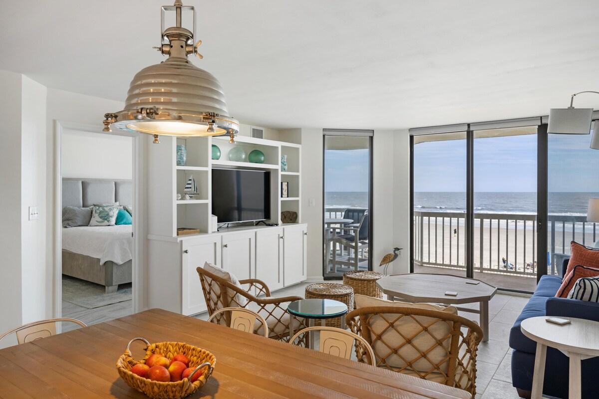 A light-filled living area features large windows with ocean views. A dining table with a fruit basket sits in the foreground, while a comfortable blue sofa and armchairs surround a coffee table. A decorative shelving unit displays books and a television.