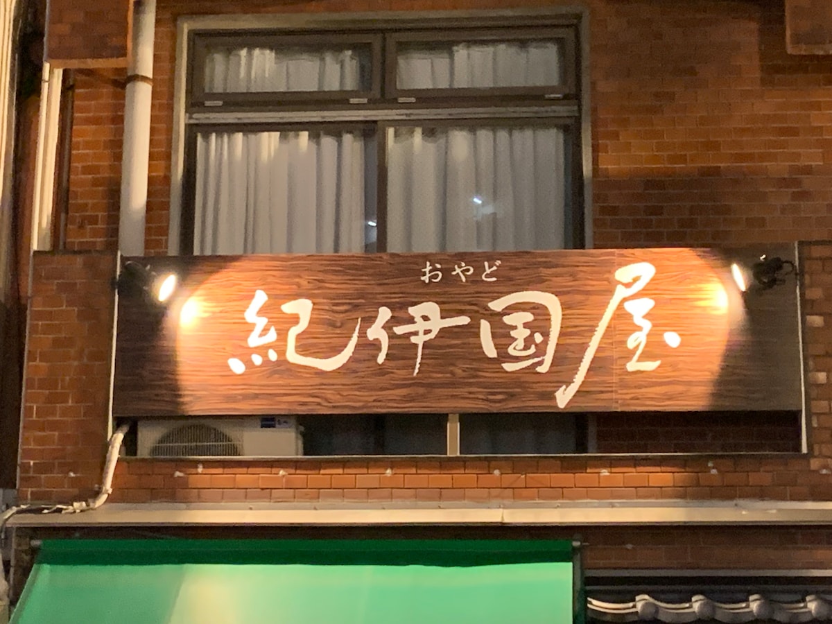 A wooden sign displaying stylized Japanese characters is highlighted by two spotlights. The sign is mounted above a storefront with a green awning. Soft light illuminates the area, enhancing the welcoming presence of the establishment.