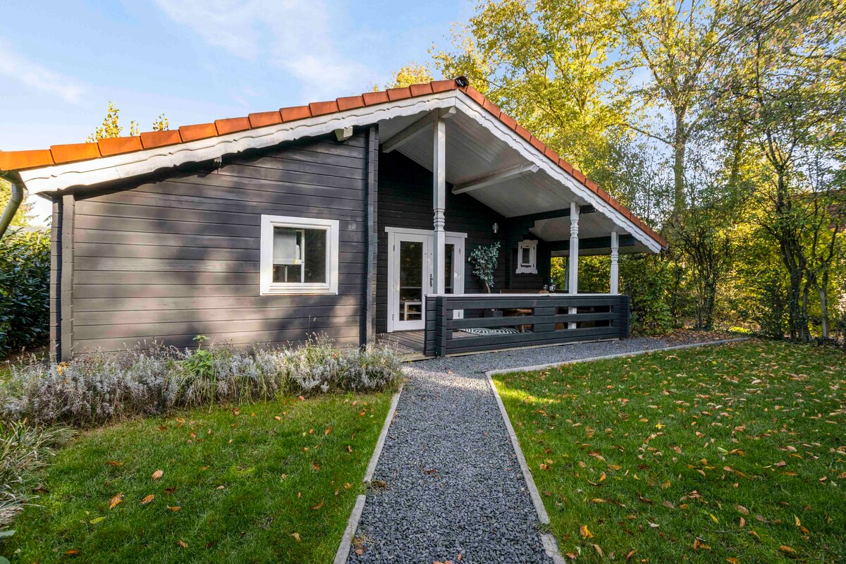 A wooden holiday home features a sloped roof with a distinctive red tile. The entrance is framed by a spacious porch, which includes outdoor seating. A gravel pathway leads from the lush green lawn to the front door, surrounded by trees and flowering shrubs.