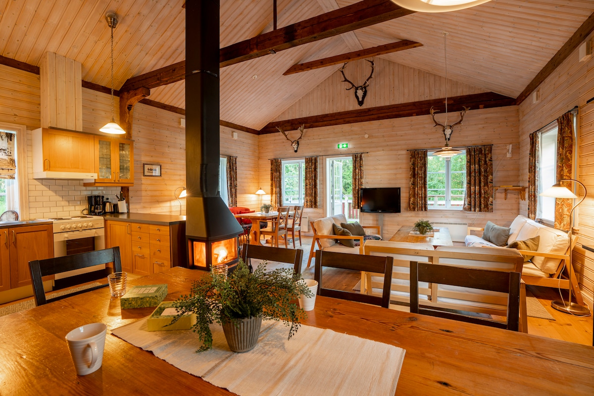 The inviting communal space features high wooden ceilings and a warm fireplace, surrounded by large windows allowing natural light. A dining table is positioned centrally, with a cozy seating area visible in the background, showcasing comfortable furnishings and a television.