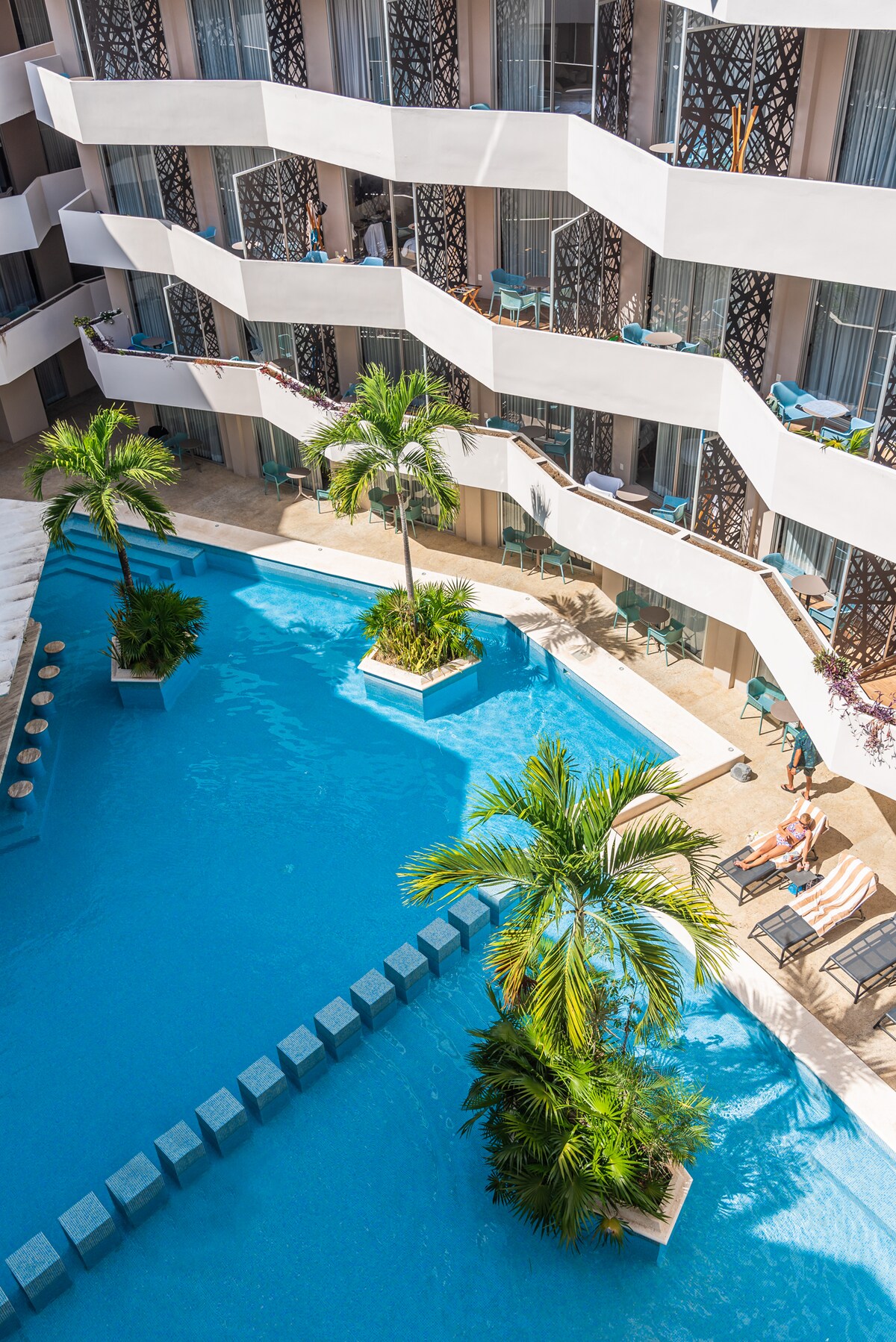 A pool area is visible, featuring clear blue water surrounded by palm trees and lounge seating. The architecture displays multiple levels with balconies adorned with teal furniture, creating a relaxed atmosphere for guests to enjoy the outdoor space.