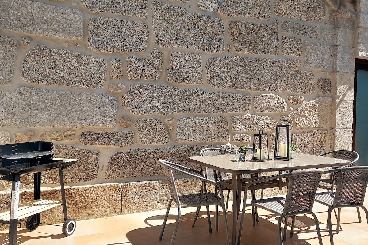 A spacious outdoor dining area is visible, featuring a table surrounded by several chairs. A charcoal grill is positioned nearby, while the backdrop consists of a rustic stone wall, adding character to the space.