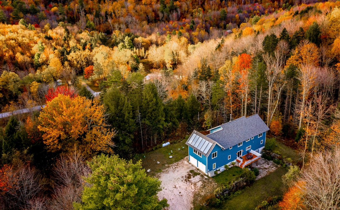 An aerial view showcases a spacious blue house surrounded by vibrant autumn foliage. Shades of orange, yellow, and green trees create a colorful backdrop, while a gravel area is visible around the property. The serene landscape reflects the tranquility of the surrounding nature.