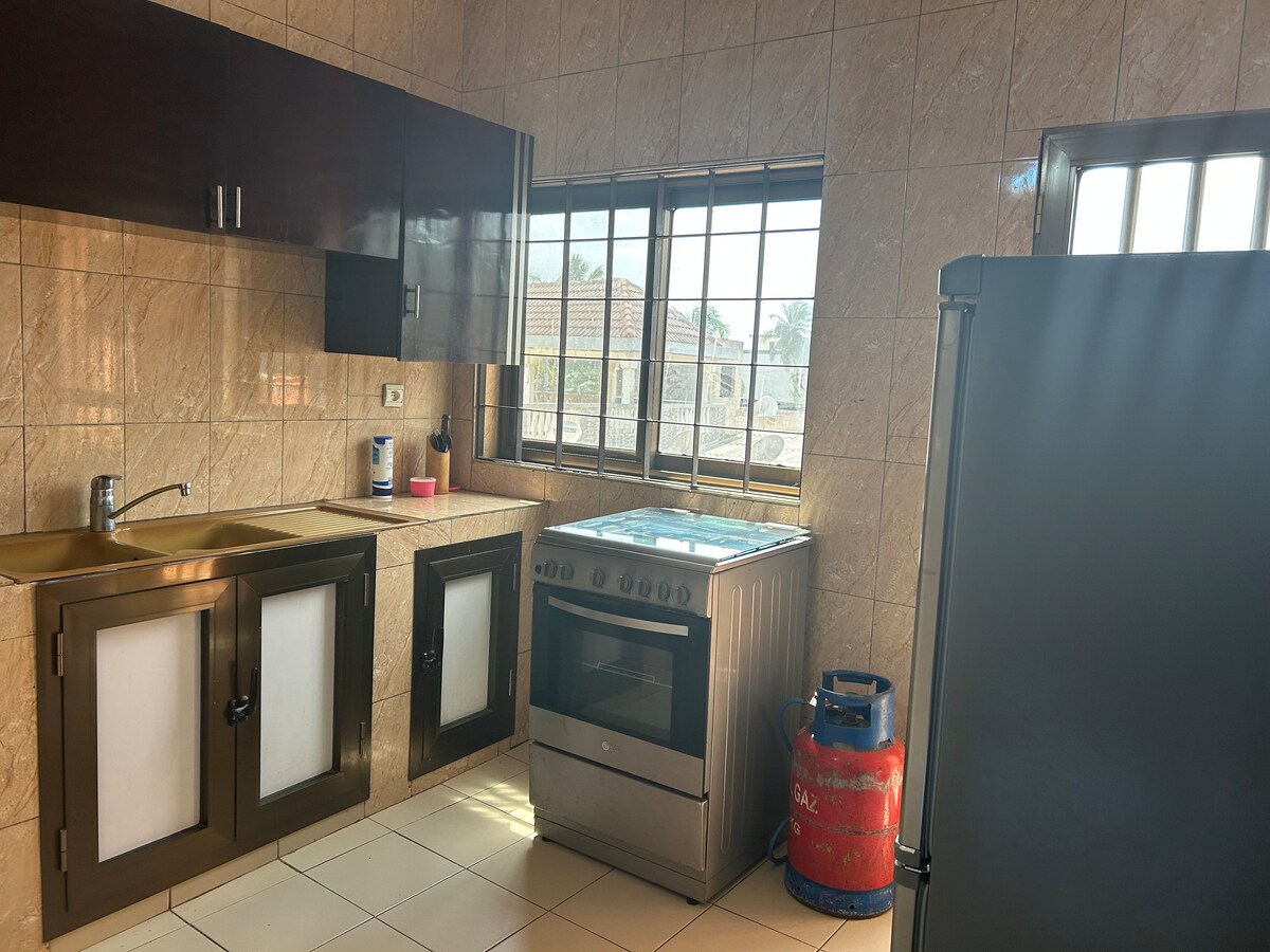 A well-equipped kitchen is visible, featuring light-colored tiled walls and a combination of dark cabinets. A gas stove is positioned beside a refrigerator. Natural light enters through large windows, illuminating the workspace with a warmth that enhances the functional layout.