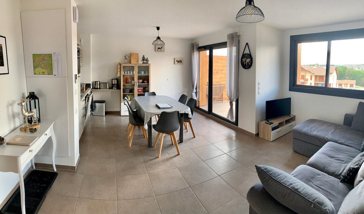 A spacious living area features a large gray sectional sofa and a dining table surrounded by six modern chairs. A sideboard and an entertainment unit are present. Large windows allow natural light to fill the room, offering views of the surrounding outdoor area.