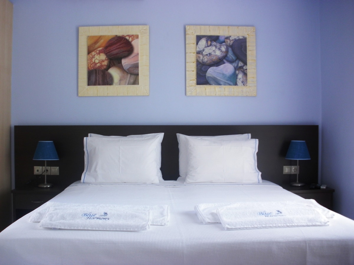A comfortable bedroom features a spacious bed with white linens and two decorative pillows. Flanking the bed are two bedside tables with blue lamps. Colorful artwork adorns the wall above the bed, adding a touch of personality to the room.