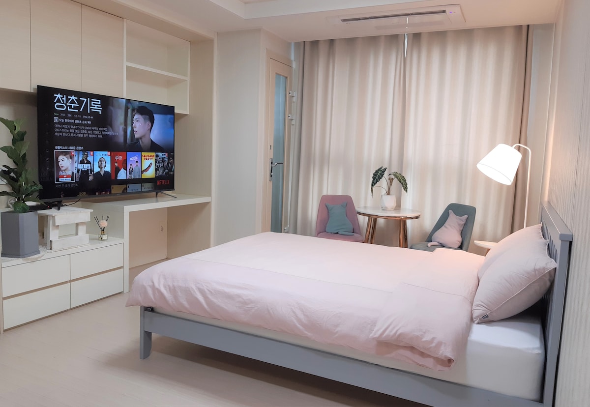 A cozy bedroom is arranged with a spacious bed covered in light pink bedding. A large flat-screen TV is mounted on a wall unit, displaying streaming content. Natural light filters through sheer curtains, complementing a small table with two chairs and a decorative plant.