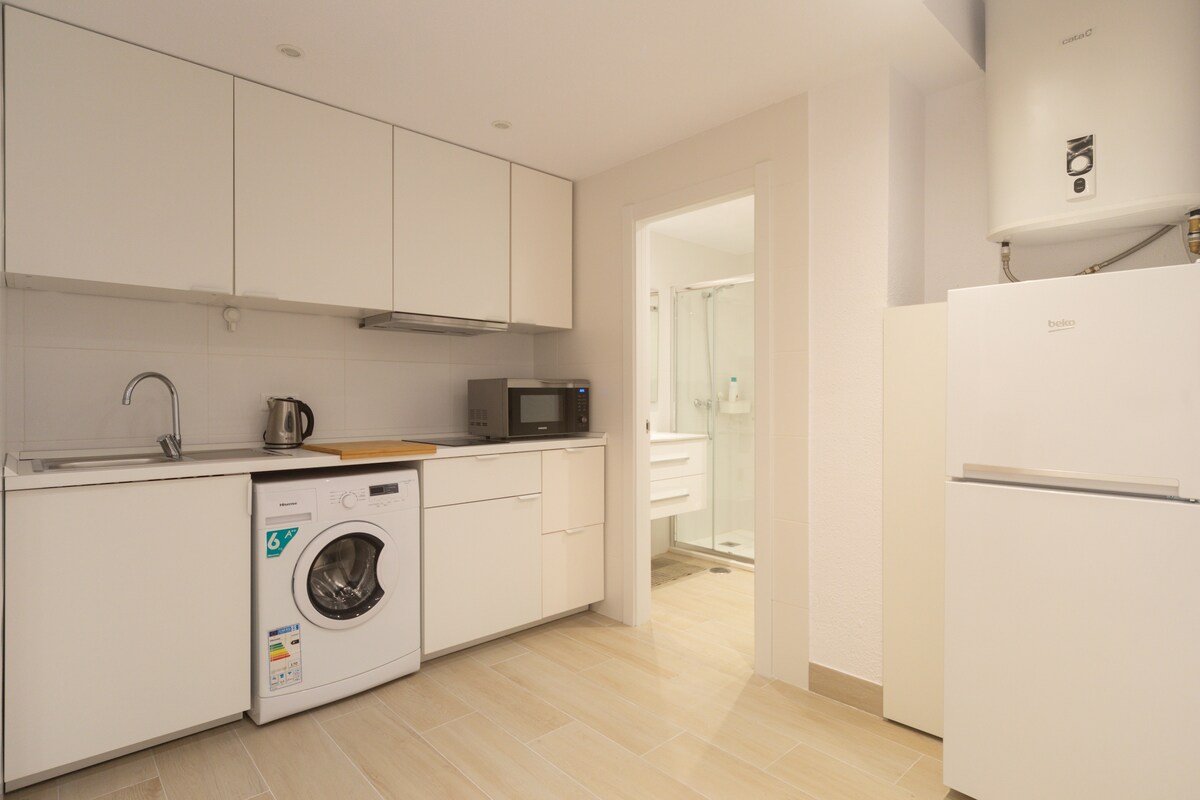 A modern kitchen space is equipped with essential appliances, including a washing machine, microwave, and refrigerator. Neutral cabinetry complements the light-colored tiled flooring, while a door leads to an adjacent bathroom. Natural light enhances the bright, clean atmosphere of the area.