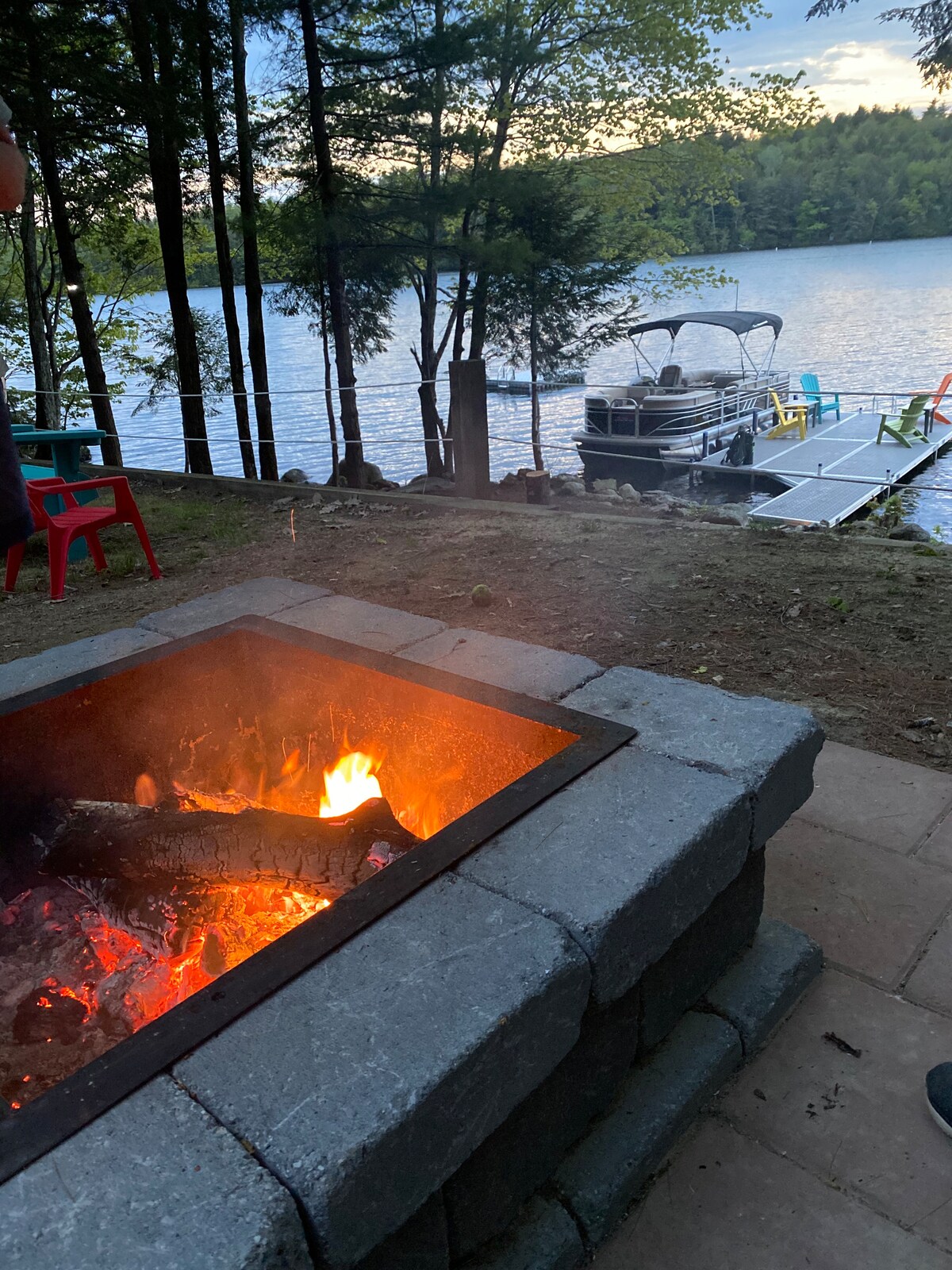 A fire pit constructed from stone blocks is seen with glowing flames and burning wood. In the background, the serene lake reflects the evening sky, with a private dock visible featuring seating. Surrounding trees provide privacy, enhancing the tranquil atmosphere.