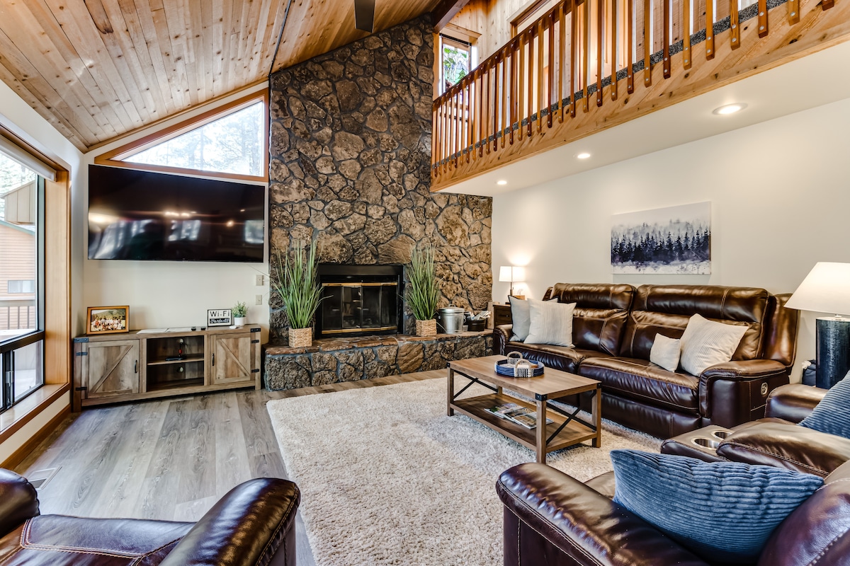 The living room features a large, stone fireplace as a focal point, surrounded by comfortable leather seating. A flat-screen TV is mounted on the wall, and natural light enters through large windows. A wooden ceiling adds warmth, while a light rug enhances the spacious feel.