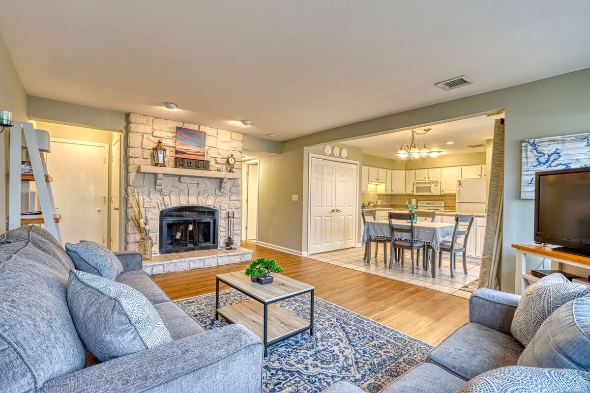 A spacious living area features a cozy fireplace framed by a decorative stone façade. Soft seating, including two plush sofas, is arranged around a central coffee table. The open layout connects to a well-lit dining area with a table set for six, enhancing the inviting atmosphere.