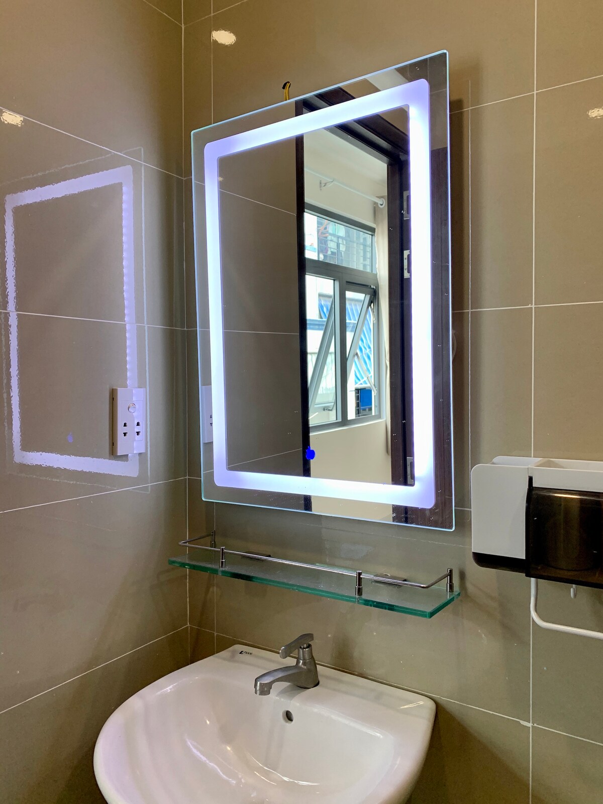 The well-maintained bathroom features modern fixtures, including a sleek illuminated mirror above a compact sink. A glass shelf is mounted underneath the mirror, providing space for toiletries. Natural light enters through a nearby window, enhancing the bright and clean atmosphere of the space.
