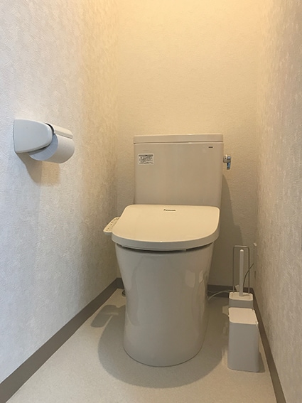 A clean and modern bathroom setup is displayed, featuring a white toilet against a light-colored wall. A toilet paper holder is mounted nearby. The floor is finished with a light-colored material, contributing to a simple and functional space.