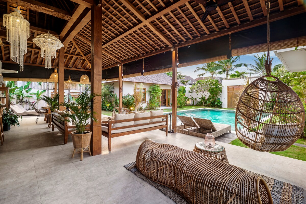 An open living area is showcased, featuring a blend of natural materials and modern design. Comfortable seating options include a large daybed and rattan furniture, surrounded by tropical greenery. The space offers direct views of the pool area, enhancing the inviting atmosphere.