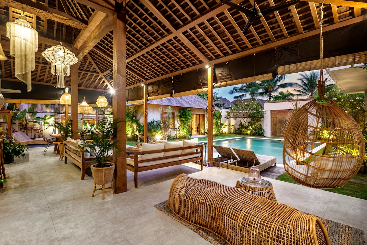 An expansive open living area is surrounded by lush greenery, featuring a large inviting pool visible in the background. Comfortable seating arrangements include a spacious sofa and unique hanging chair, with warm lighting enhancing the tropical ambiance of the villa.