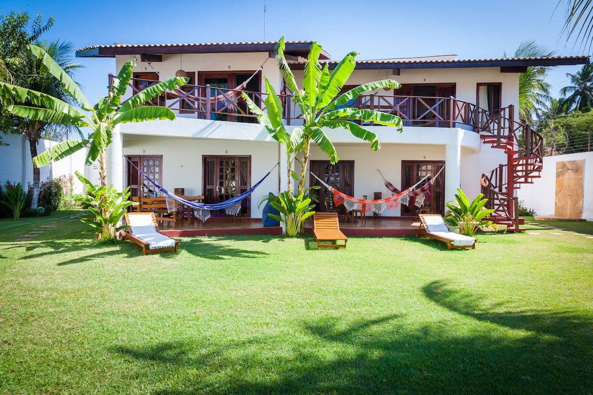 An inviting exterior view showcases a two-story building surrounded by lush greenery. Several hammocks are positioned between palm trees, while sun loungers are arranged on the well-maintained lawn. The balcony features a staircase, enhancing access to the upper level.