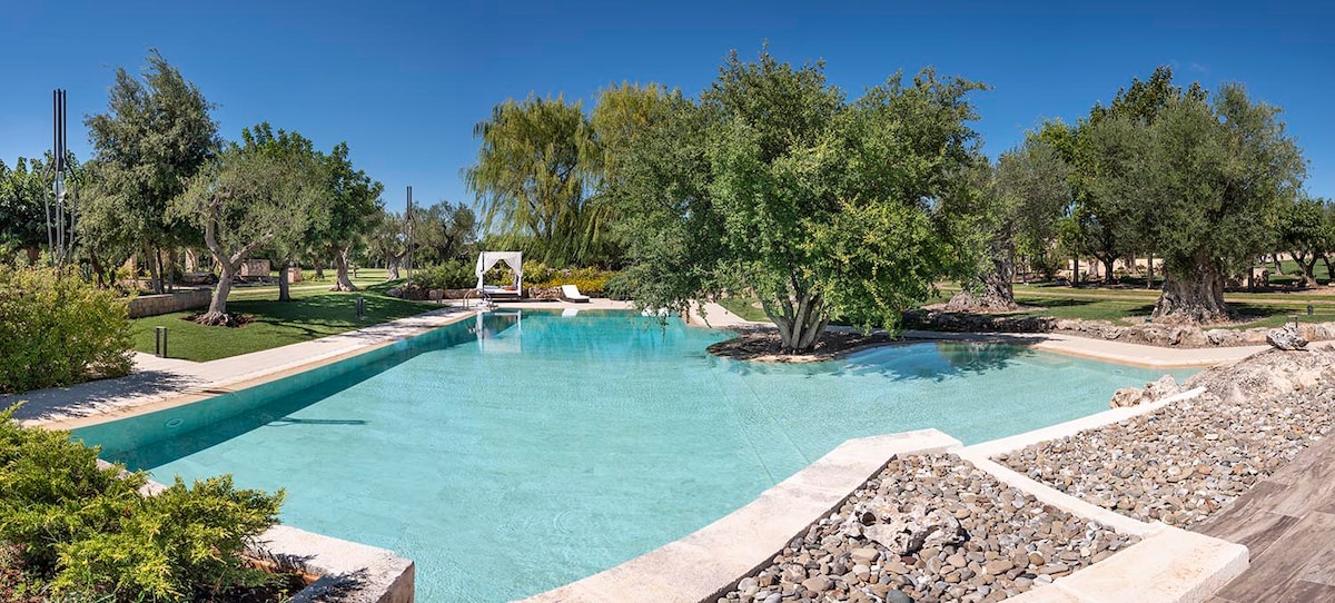 An expansive outdoor pool is surrounded by lush greenery and mature trees. The clear water reflects the blue sky, while a shaded seating area is visible nearby. Natural stone accents enhance the serene landscape, providing a tranquil setting for relaxation.