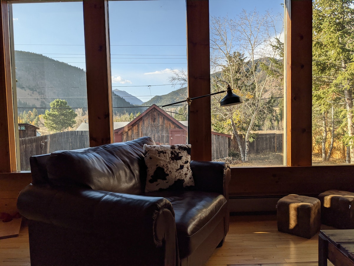 A cozy seating area features a leather chair accented with a decorative pillow. Large windows frame stunning mountain views, allowing natural light to fill the space. The rustic atmosphere is complemented by natural wood finishes and soft furnishings.