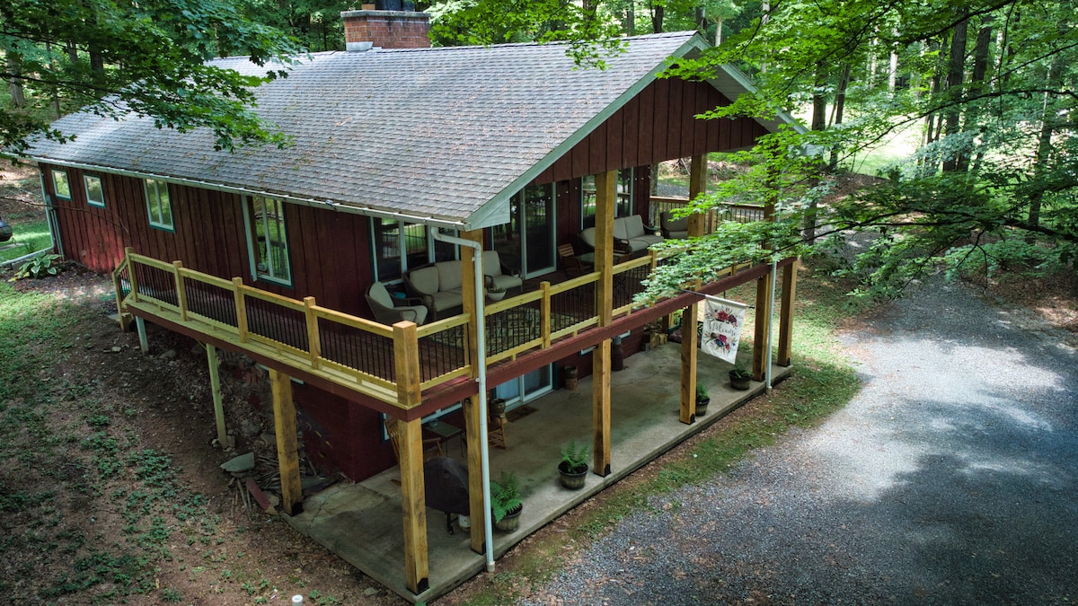 The exterior of Cranberry Cottage showcases a welcoming wooden structure with a red facade, surrounded by lush greenery. A spacious covered porch offers ample seating, inviting relaxation in a tranquil wooded setting. A gravel driveway leads to the home's entrance, enhancing its secluded charm.