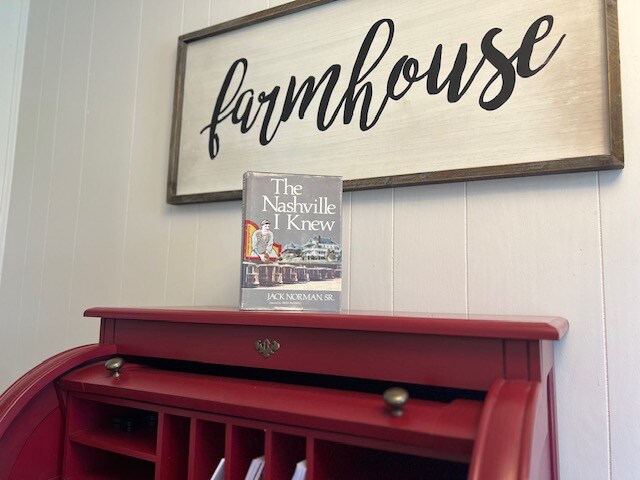A rustic red writing desk sits beneath a wooden sign that reads 'farmhouse.' The desk displays a book titled 'The Nashville I Knew,' adding a touch of local culture. Light colors of the wall enhance the warm and inviting setting.