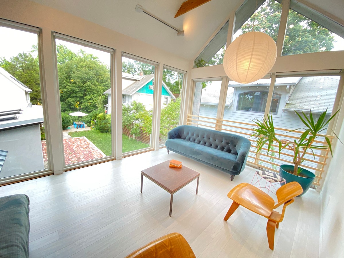 The living area features a large window wall, allowing natural light to fill the space. A plush blue sofa and a wooden chair are situated around a small table. A pendant light hangs from the ceiling, and indoor plants add a touch of greenery.
