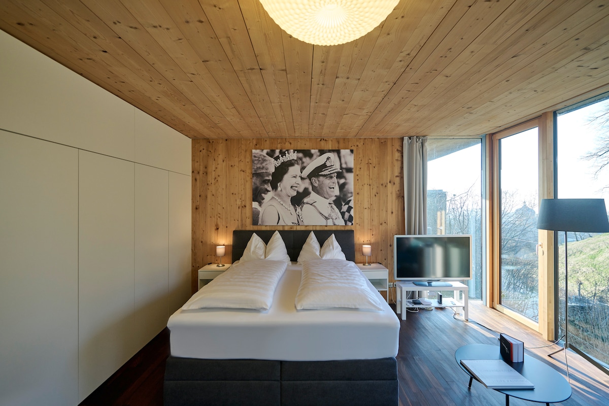 A well-appointed sleeping area is showcased with a spacious bed featuring crisp white linens and decorative pillows. A large artwork is displayed on the wood-paneled wall. Natural light floods the room through expansive windows, complemented by a modern TV and a sleek lamp.