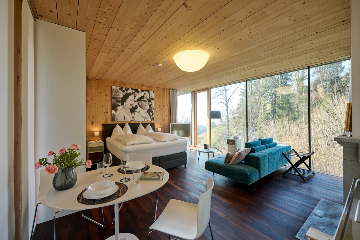 A spacious living and sleeping area features a large bed framed by wood paneling. A modern turquoise sofa complements a round dining table set for two. Extensive floor-to-ceiling windows offer a view of the surrounding nature, enhancing the room's connection to the outdoors.