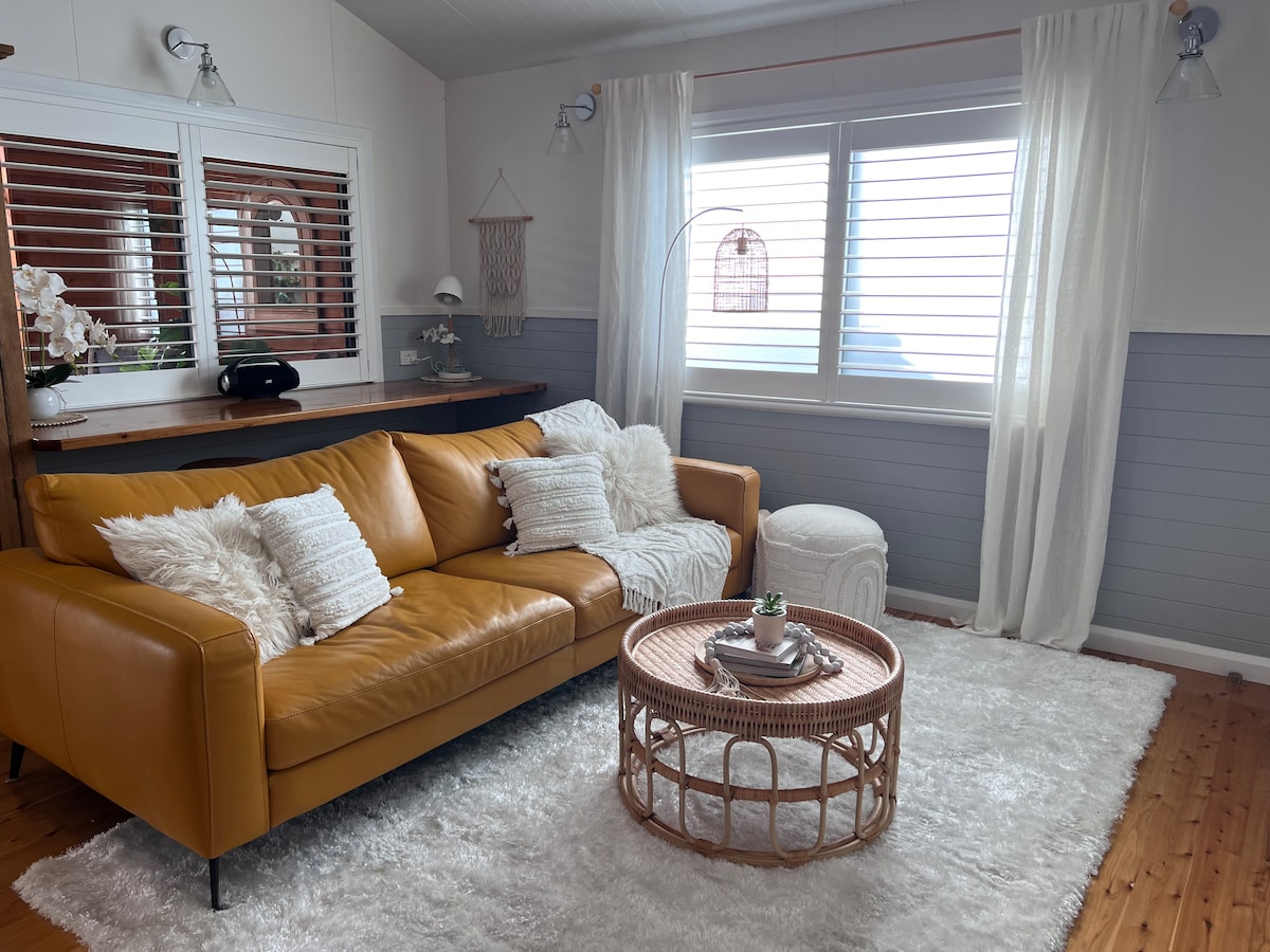 A cozy living room is presented with a sleek leather sofa adorned with decorative cushions. A round wicker coffee table sits atop a plush area rug, while light filters through a window draped with sheer curtains. A calming atmosphere is created by the soft furnishings.