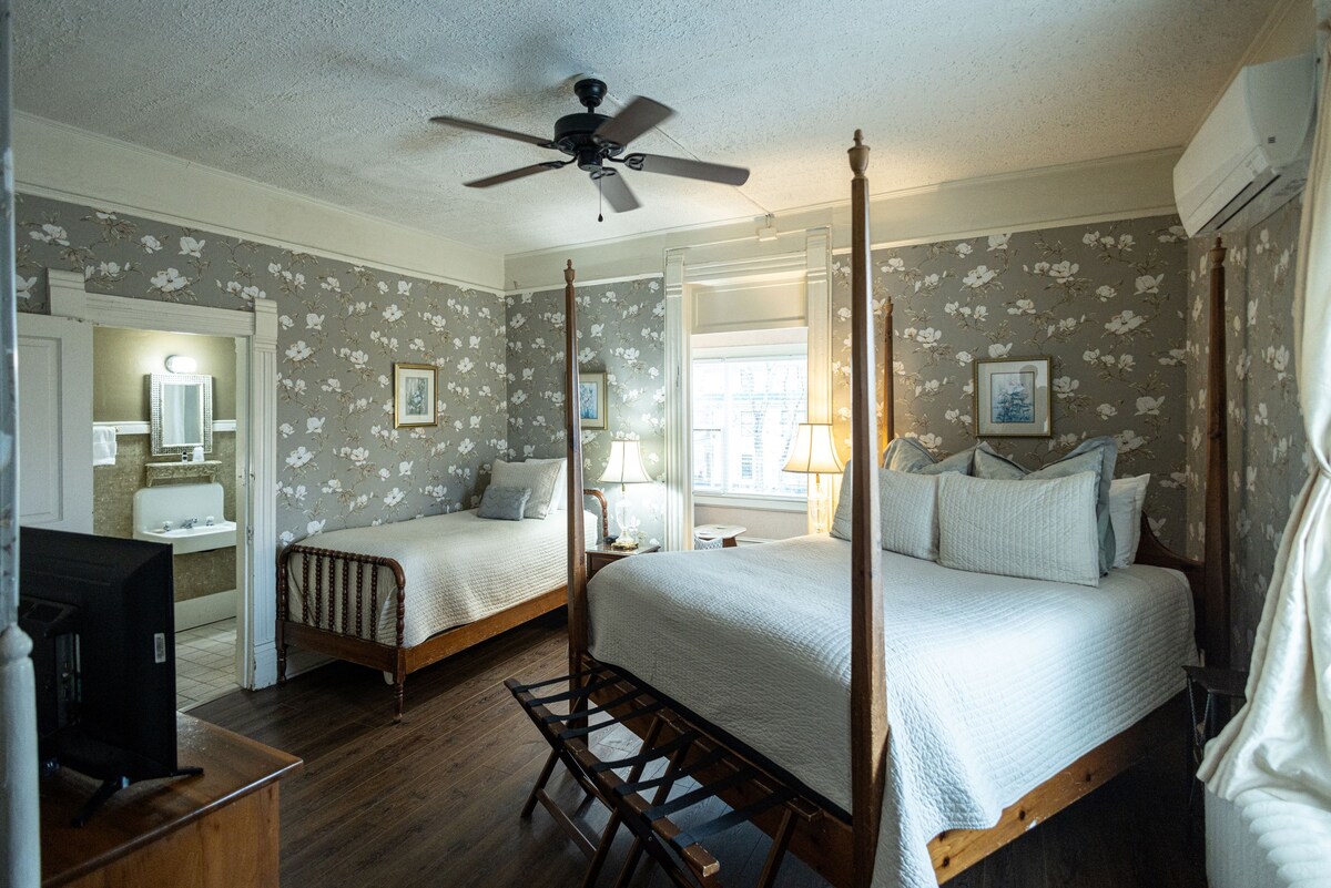 The spacious room features a queen bed and a twin bed, with wooden bed frames. A ceiling fan and lamps provide illumination. A small TV is positioned on a wooden dresser. A doorway leads to an adjacent bathroom with visible light tiles.