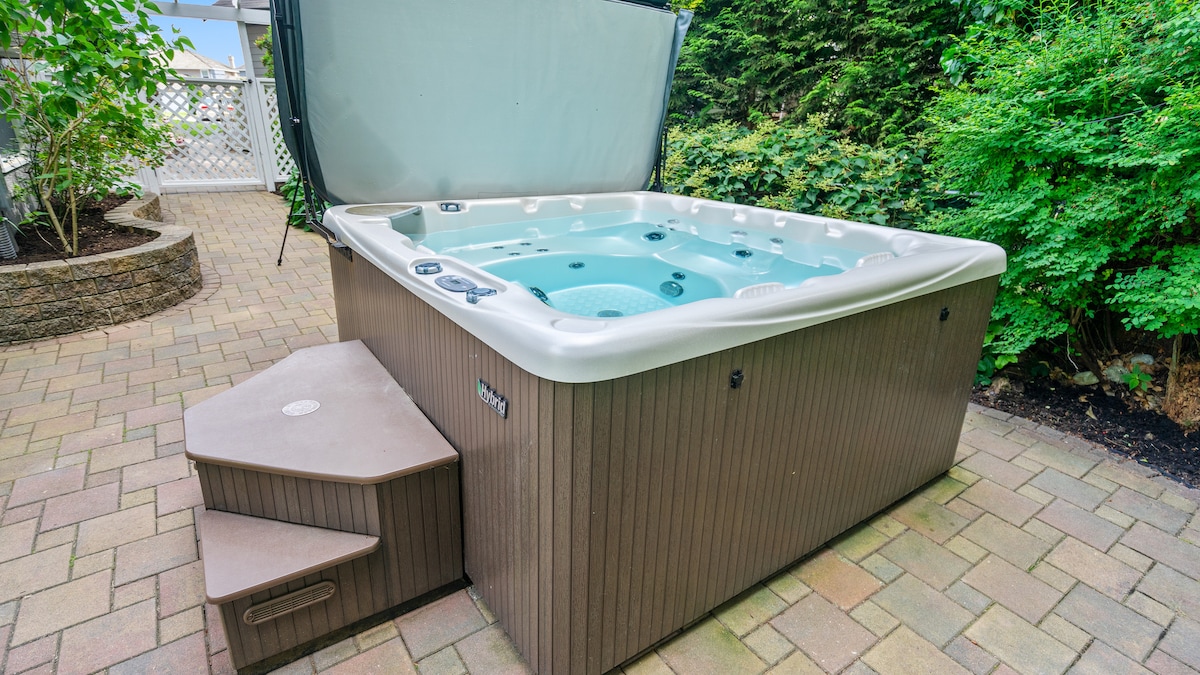 A private hot tub is situated on a stone-paved patio surrounded by lush greenery. The hot tub features a sleek exterior with steps for easy access. Clear water invites relaxation, while the structure is covered for weather protection.