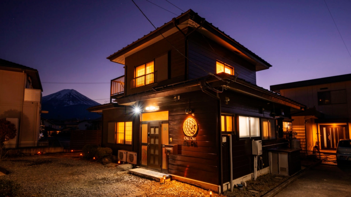 The exterior of the villa is illuminated at dusk, showcasing a traditional design with warm lights glowing from the windows. Mt. Fuji is visible in the background, providing a serene backdrop to the charming structure.