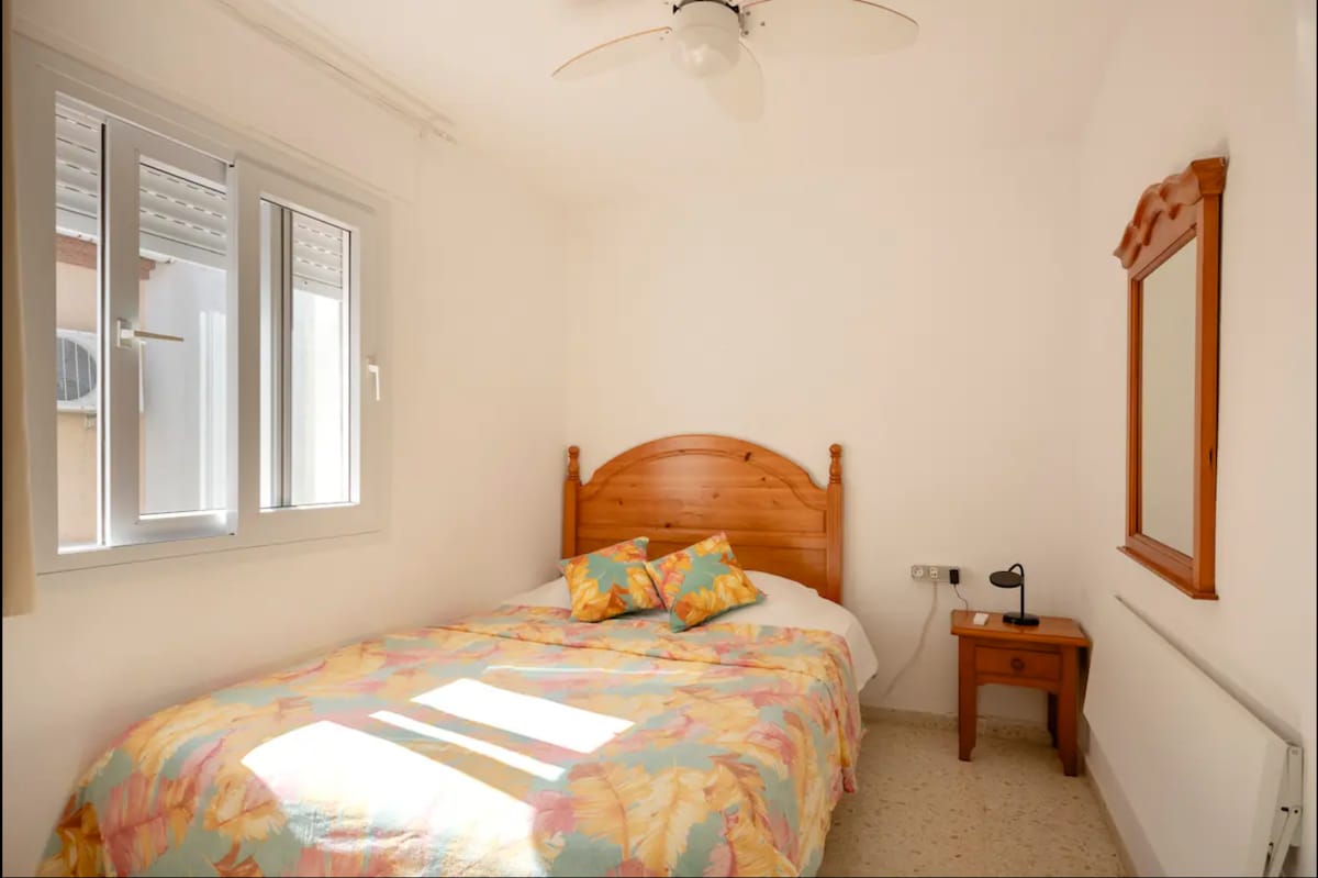 A bright room features a spacious bed with a colorful quilt and two decorative pillows. A small nightstand with a lamp is positioned beside the bed, while a wooden mirror adds a practical touch. Natural light enters through a window with a soft curtain.