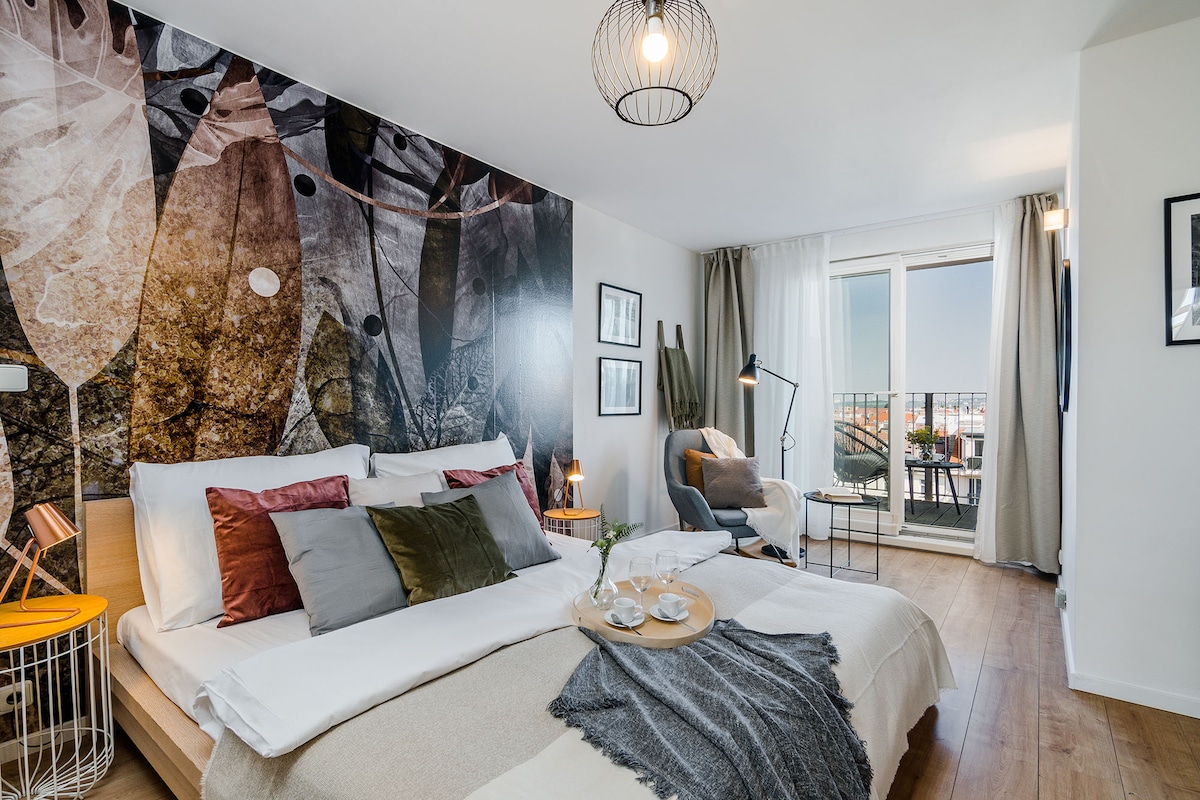 A modern bedroom features a large bed with decorative pillows in neutral tones. A wall adorned with artistic textures creates visual interest. Nearby, a cozy seating area includes a chair and side table, with natural light filtering through the balcony doors onto a small outdoor space.