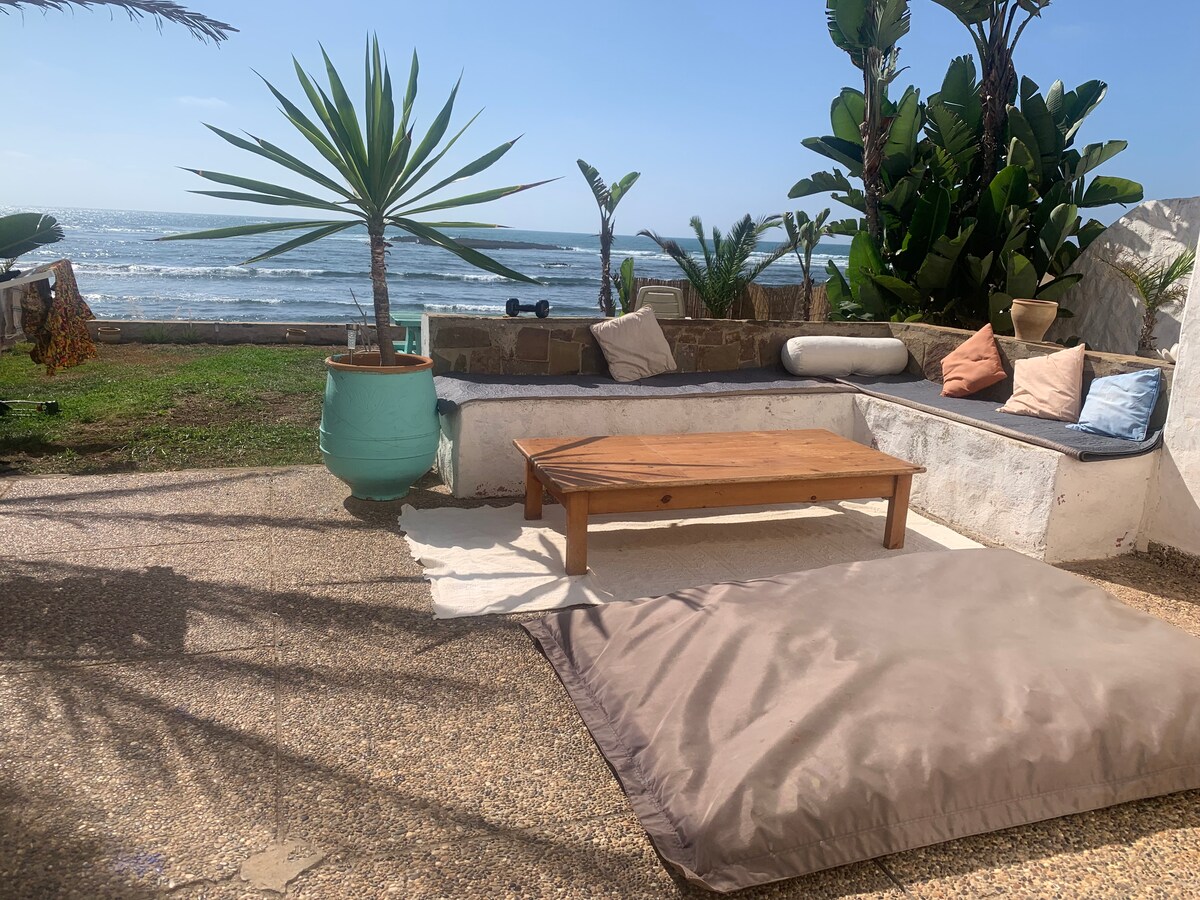 An outdoor lounging area is displayed, featuring a low wooden table surrounded by a spacious cushion. Green plants are positioned nearby, and the ocean can be seen in the background, providing a serene coastal setting.