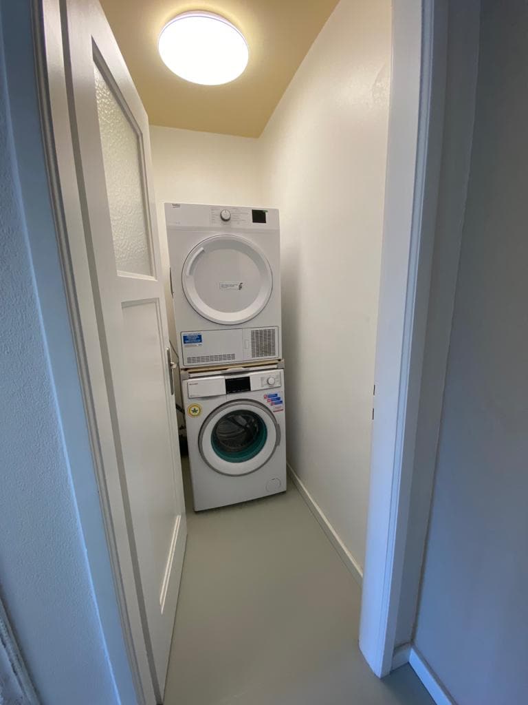 A compact laundry area is visible, featuring a stacked washer and dryer unit. The appliances are positioned against a white wall, with a light fixture above them illuminating the space. The floor is a smooth, light grey, contributing to the clean appearance.