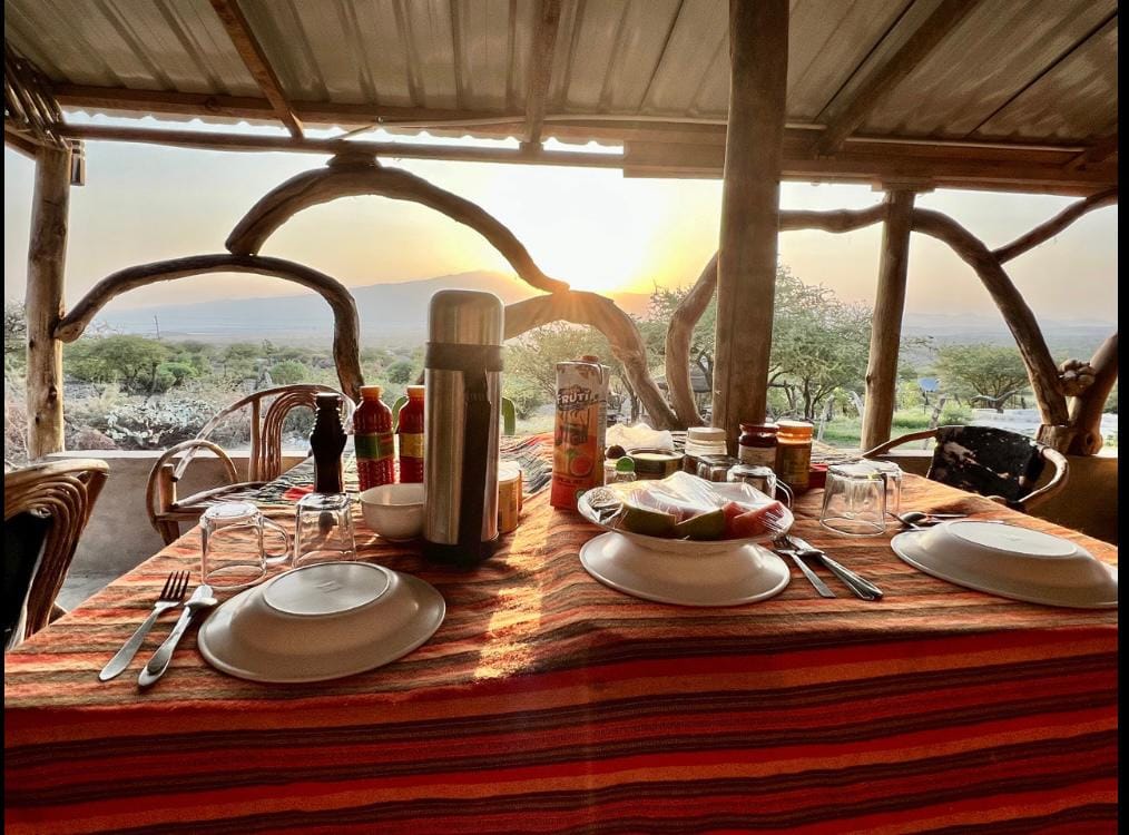A dining table set for two is displayed, featuring plates, glasses, cutlery, and various condiments. A scenic view is visible in the background, with the sun setting over a vast landscape, creating a warm glow through the ample outdoor setting.