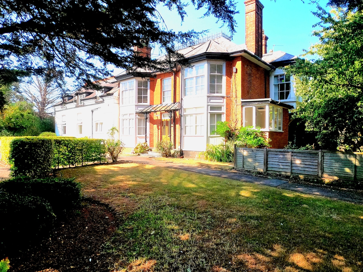 A historic Georgian-style building is surrounded by well-kept greenery. The façade features large windows and a covered entrance, offering a welcoming appearance. A manicured lawn leads up to the building, providing a spacious outdoor area that blends with the natural surroundings.