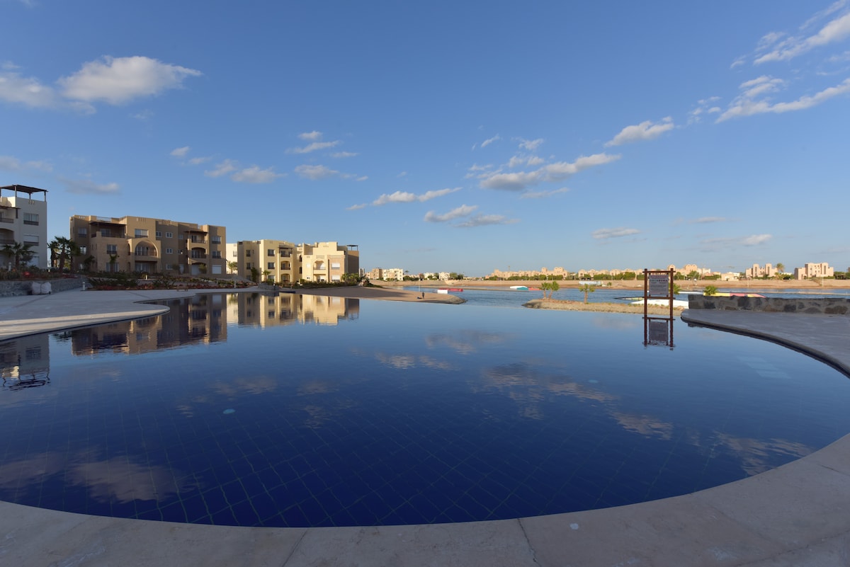The image captures a serene infinity pool reflecting the clear blue sky and surrounding buildings. The calm waters blend seamlessly with the horizon, while a gentle landscape of greenery and distant structures creates a tranquil atmosphere. The pool area appears spacious and well-maintained.