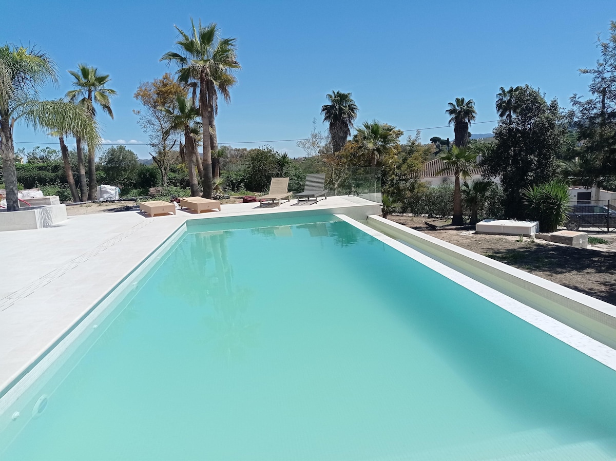 An infinity pool is framed by palm trees and well-maintained greenery. Sun loungers are positioned nearby, offering a relaxing space. The pool's water appears calm and inviting, reflecting the clear blue sky above. The surrounding area consists of landscaped gardens and open space.