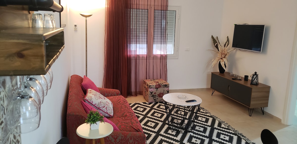 A bright living area is furnished with a red patterned sofa and a colorful ottoman. A small round table sits on a geometric rug, while a flat-screen television is mounted on the wall. Large windows are adorned with sheer curtains, allowing natural light to flow in.