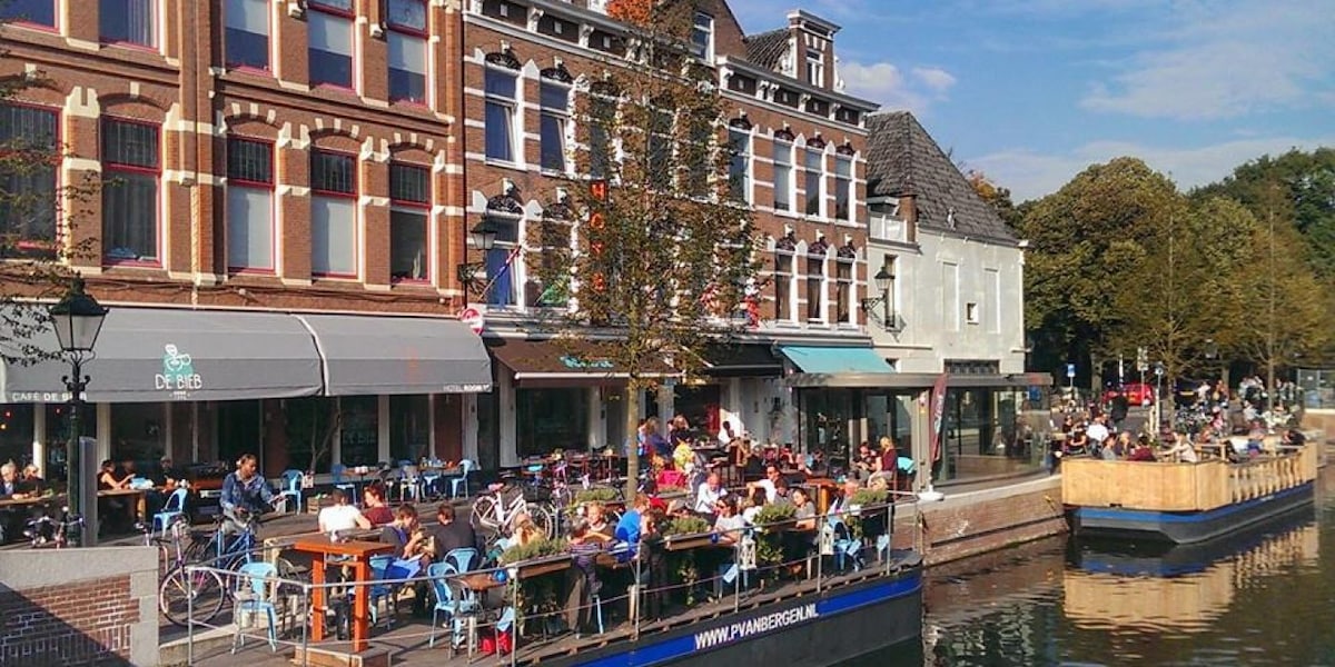 A lively waterfront scene is observed with a busy terrace filled with patrons seated at blue chairs. Trees provide greenery along the riverside, while charming buildings rise in the background, showcasing a mix of architectural styles typical of the area.