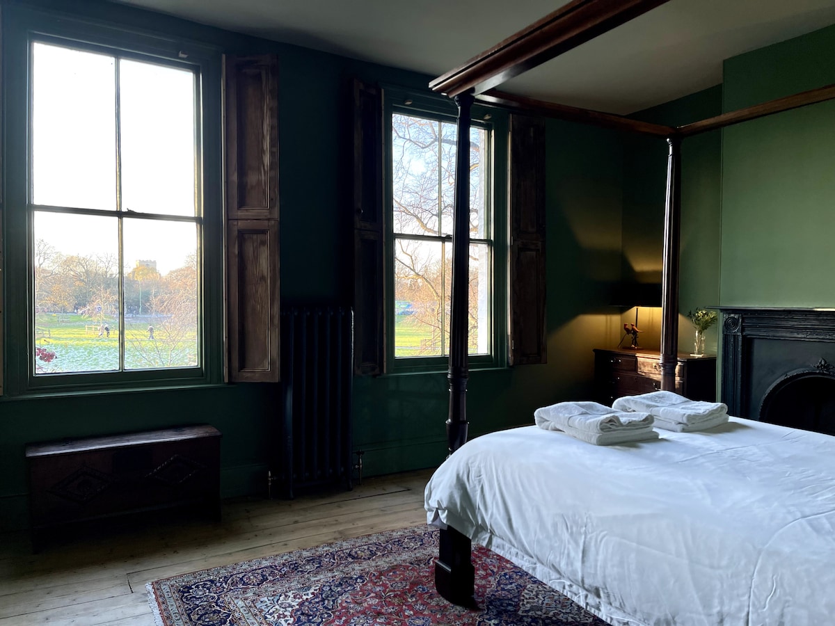 The bedroom showcases a four-poster bed with fresh white linens, positioned by large windows offering views of Midsummer Common. The dark green walls and natural light create a peaceful ambiance, while a wooden trunk and antique dresser complement the space.